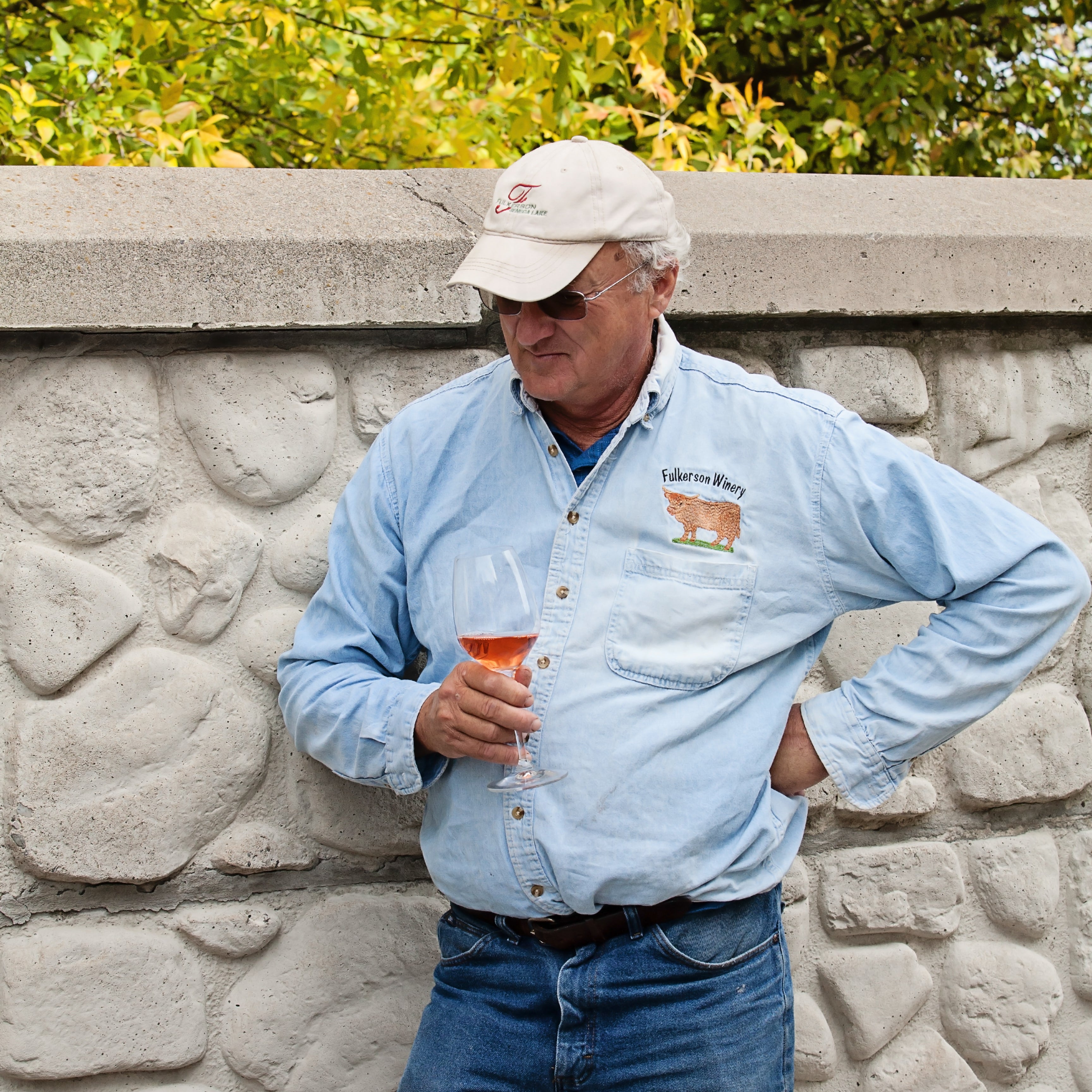 Man in a light blue shirt and cap holding a glass against a stone wall with trees in the background