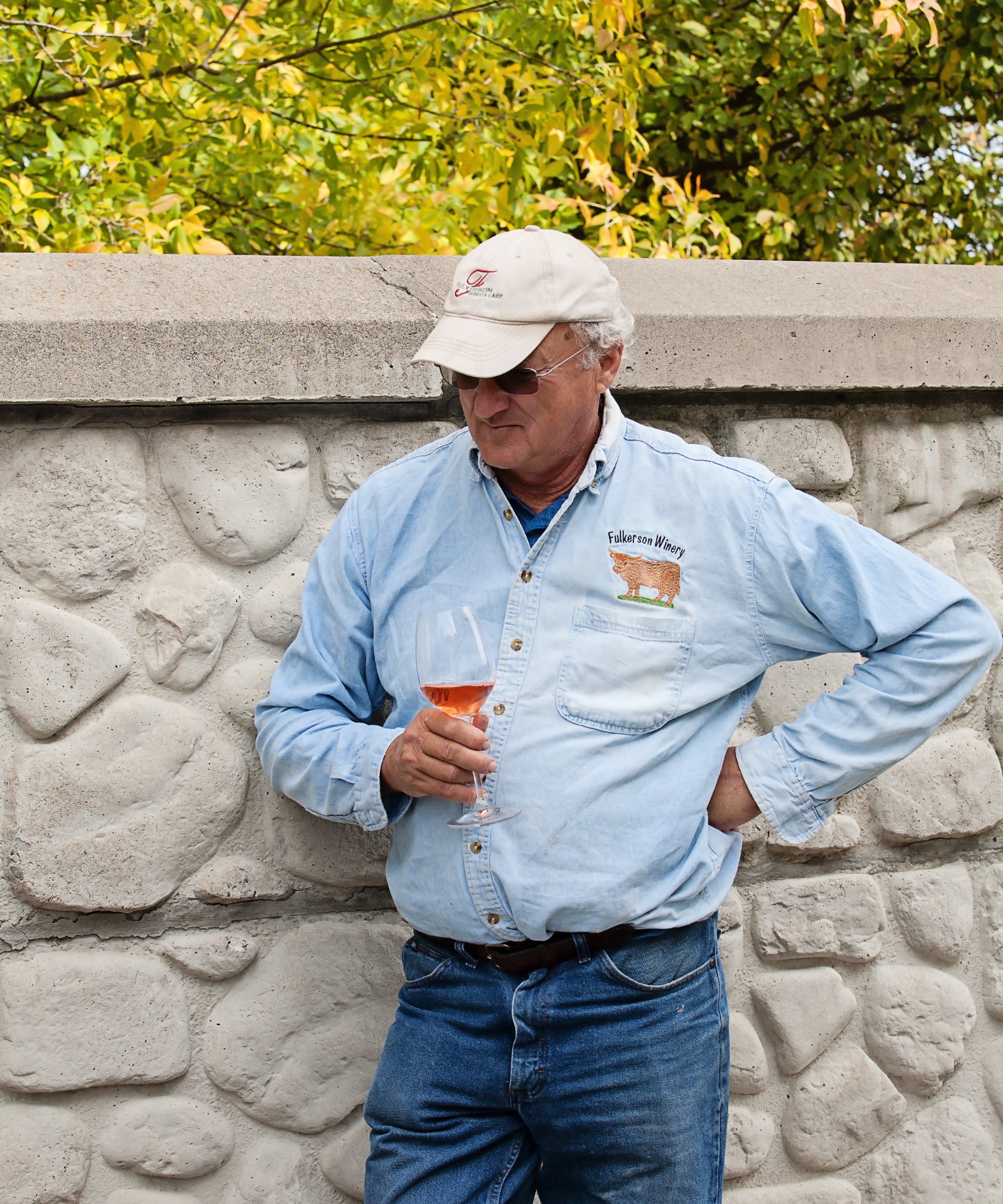 Man in a light blue shirt and cap holding a glass against a stone wall with trees in the background