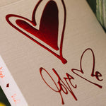Card with red heart and 'Love' text on a beige background, surrounded by green leaves.
