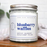 Candle jar labeled 'Blueberry Waffles' on a wooden surface with a white towel and greenery in the background.